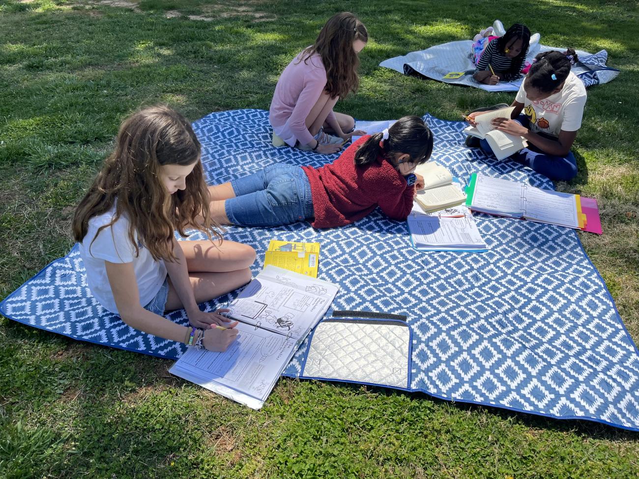 4 young girls doing schoolwork outside on a picnic blanket